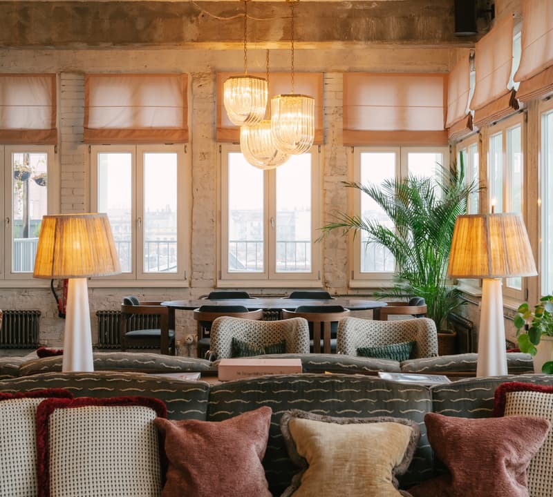 A cozy lounge area with plush pillows on sofas, two lit table lamps, hanging chandeliers, large windows with sheer curtains, and potted plants in an industrial-style interior.