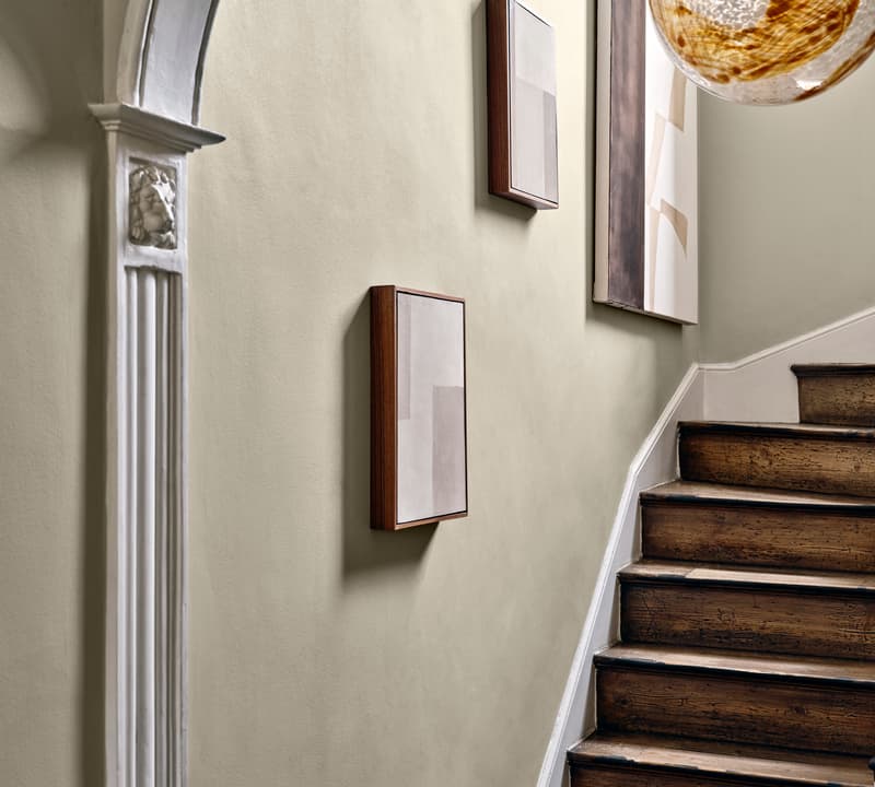Staircase with wooden steps ascends beside a light-colored wall adorned with minimalist art. An ornate archway and hanging glass light fixture enhance the elegant, contemporary setting.