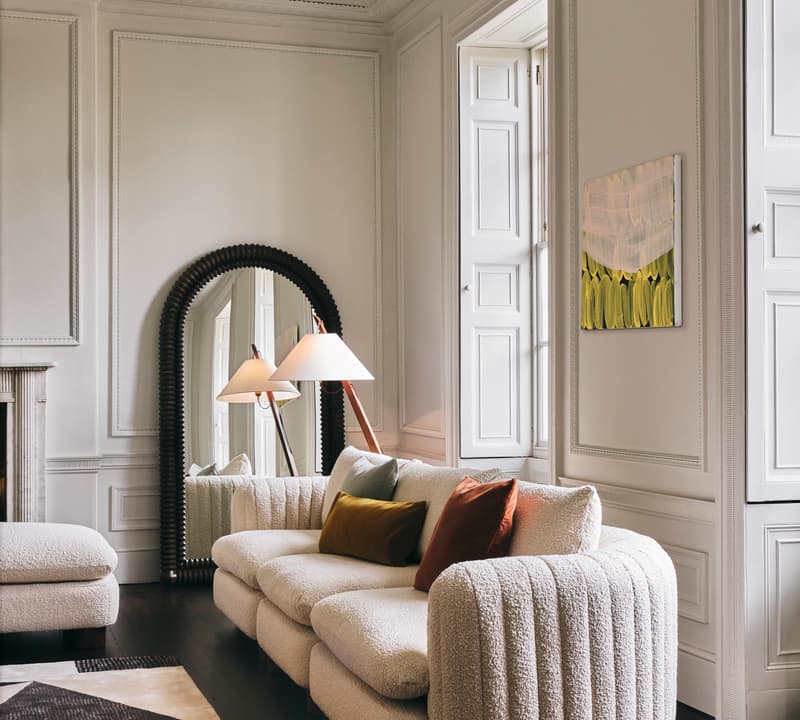 A cozy white sofa with colorful cushions stands in an elegantly decorated room with intricate white paneling, a large mirror, a floor lamp, wooden floors, and a patterned rug.
