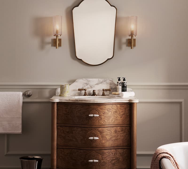 A wooden vanity with a marble countertop supports a mirror and two wall-mounted light fixtures, set in an elegant bathroom featuring a towel holder, wastebasket, and partial view of a bathtub.