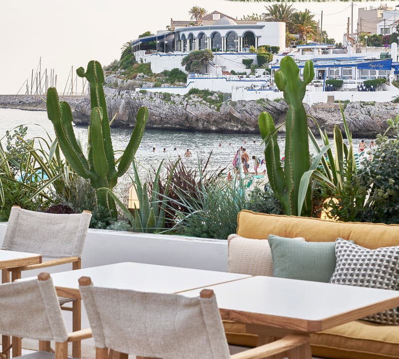 Tables and chairs arranged on a terrace are facing a seaside view with cacti, plants, beachgoers, white buildings, and boats in the background. Green-fringed umbrella partially visible above.