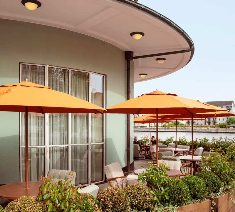 Orange umbrellas and wooden planters decorate an outdoor terrace with tables and cushioned chairs. The terrace extends from a curved green building with large windows and overlooks a waterway.