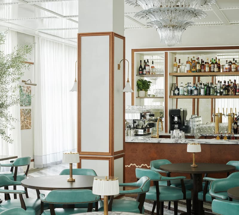 Chandelier hanging in the center of an elegant bar-café with teal chairs and round tables. A well-stocked bar, marble surfaces, a modern column, and lush greenery complete the setting.