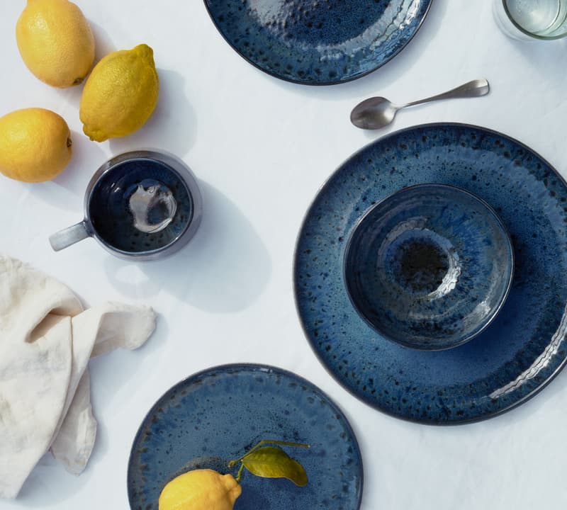 Blue ceramic plates and a mug are placed on a white tablecloth. Three lemons are arranged beside the ceramics, and a silver spoon is next to a clear glass of water.