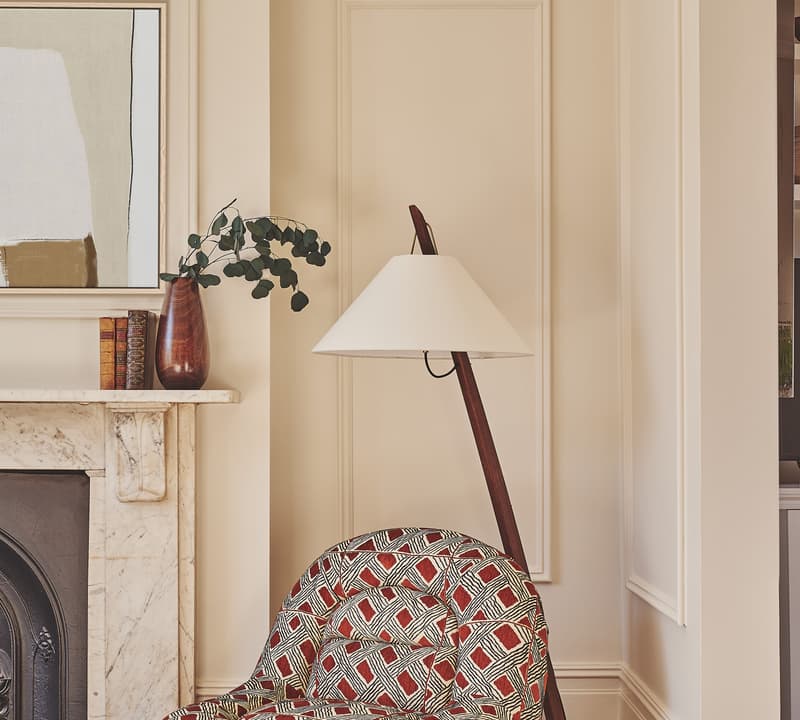 Patterned armchair sits beside a modern floor lamp in a cozy living room, near a marble fireplace adorned with a vase holding greenery and two books. An abstract painting hangs above.