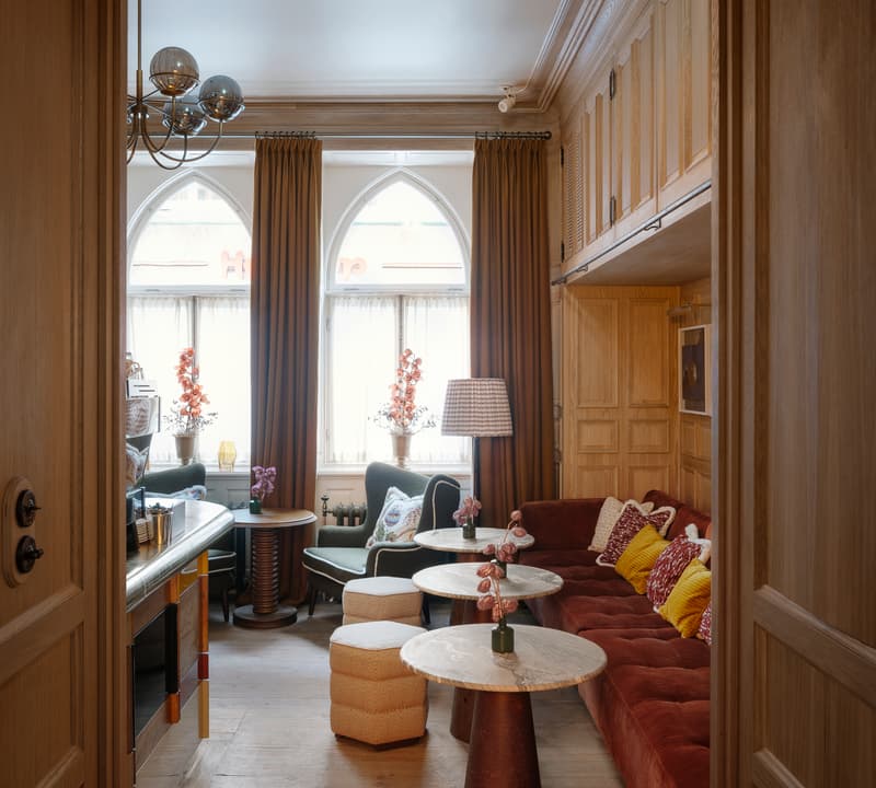 A cozy living room features a maroon sectional sofa with colorful cushions, marble tables, and green armchairs beneath arched windows with mustard-colored drapes, surrounded by warm wooden paneling.