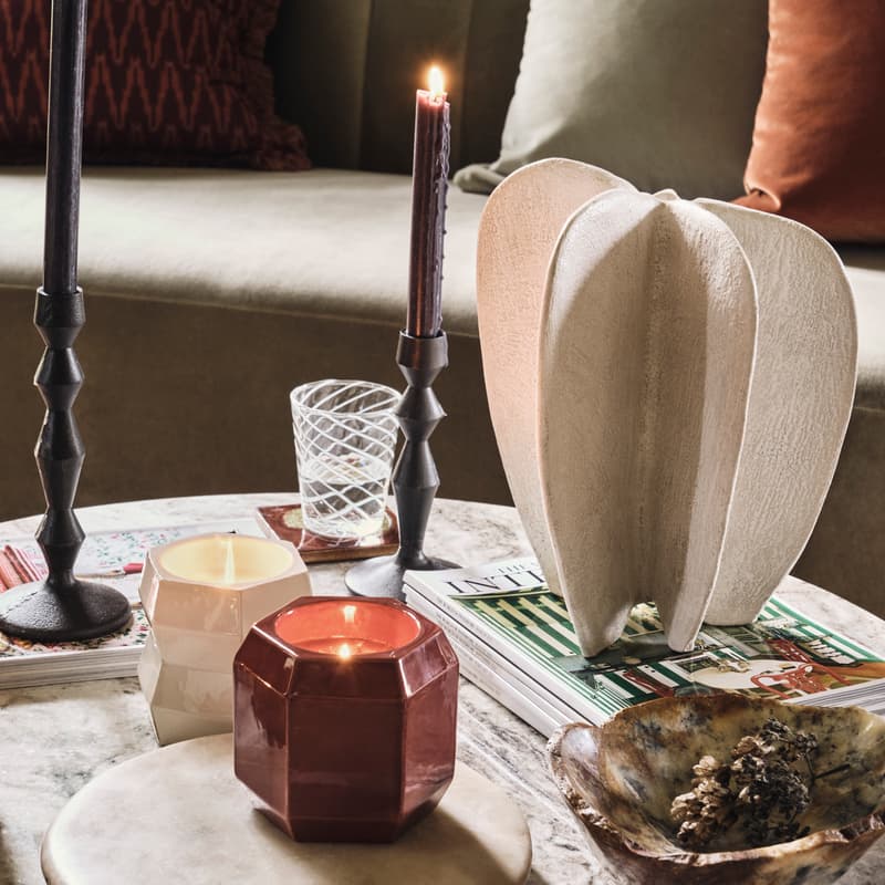 Candles and dark candlesticks are arranged on a marble table, alongside decorative items and magazines in a cozy living room with a green sofa and cushions in the background.