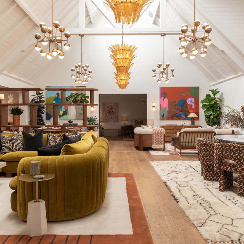 A large living room features a mustard-yellow velvet couch with patterned cushions, surrounded by modern decor, artwork, plants, wooden furniture, and hanging light fixtures under a vaulted, white ceiling.