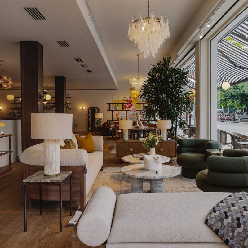 A cozy, modern showroom displays furniture, including sofas, lamps, and tables, with large glass windows revealing an outdoor patio and street view, adorned with plants and stylish decor elements.
