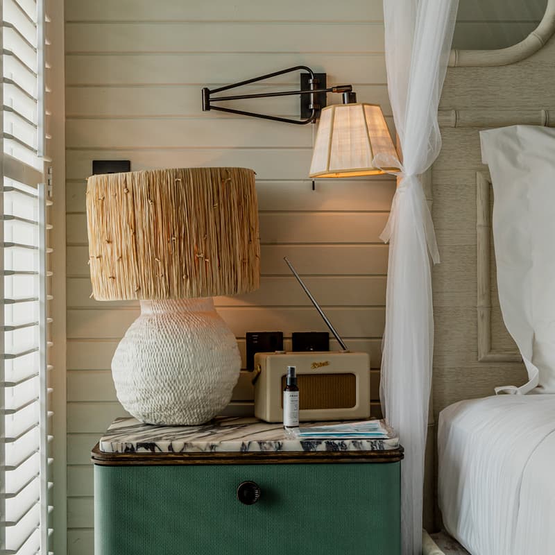 A bedside table holds a rustic lamp, a retro radio, and a small bottle in a cozy bedroom with white blinds on the window, a wall-mounted lamp, and white bedding.