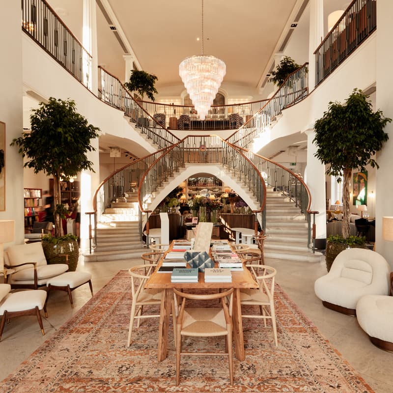 A wooden table with books and chairs sits on a rug in a luxurious room featuring a grand double staircase, large artwork, lush plants, and elegant seating.