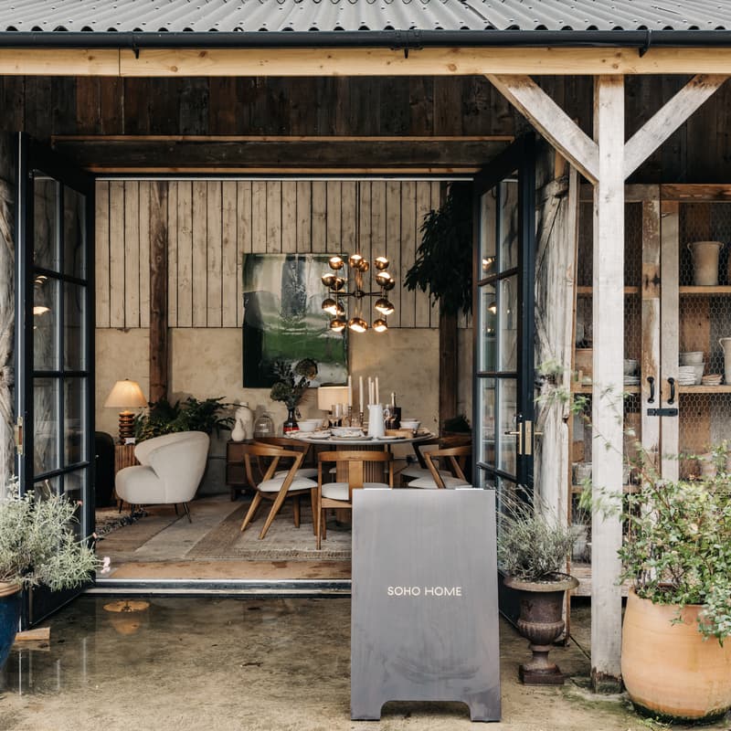A cozy showroom features a dining table setting with chairs, surrounded by rustic decor. Potted plants and pottery sit outside; a sign reads "SOHO HOME."