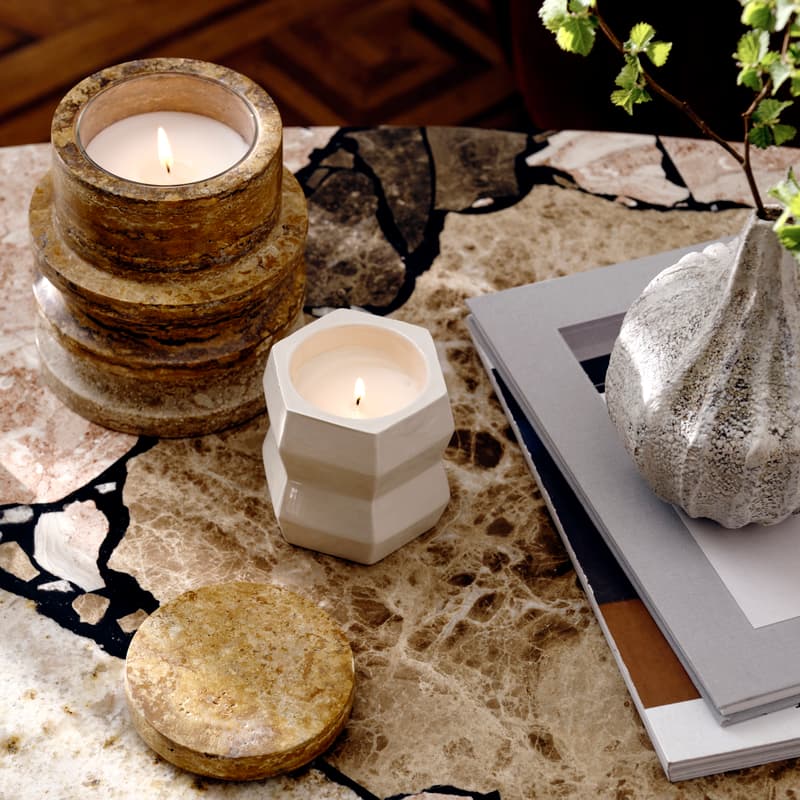 Candles burning on a marble table, one in a rustic stone holder and another in a white geometric holder. Nearby, a gold plate holds two matches, and a textured vase with greenery sits on books.