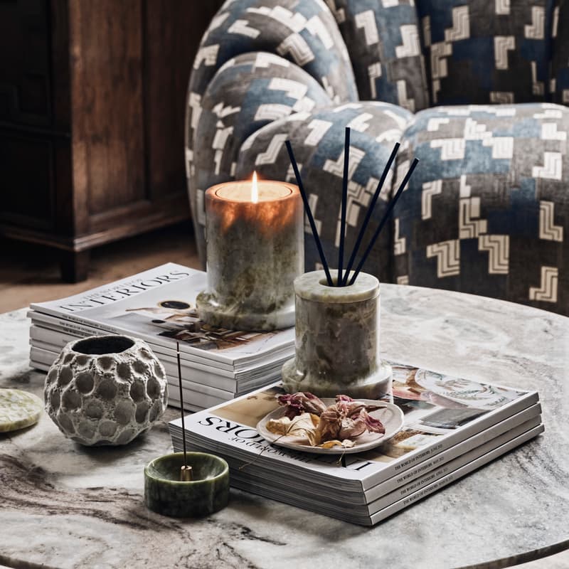 A lit candle and a reed diffuser sit on top of stacked magazines titled "INTERIORS," on a marble coffee table in a cozy living room with a patterned armchair and wooden cabinet.