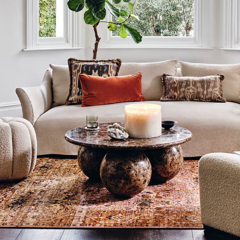 A beige sofa with decorative pillows stands in a stylish living room, surrounded by a large window, indoor plant, round ottoman, textured chairs, and a coffee table on a patterned rug.