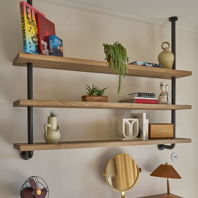 Three wooden shelves hold books, potted plants, vases, and a Marshall speaker. Below, a wooden desk with a mirror, lamp, vintage fan, book, and green stool create a cozy workspace.