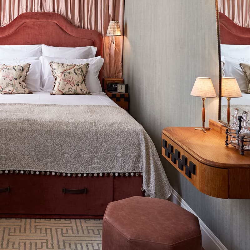 A cozy bedroom features a neatly made double bed with floral pillows and taupe bedding, flanked by pink drapes. A wooden side table with a lamp and book, “EAT DRINK NAP,” sits in front of a blue armchair. An ornate dressing table with a mirror and lamps i