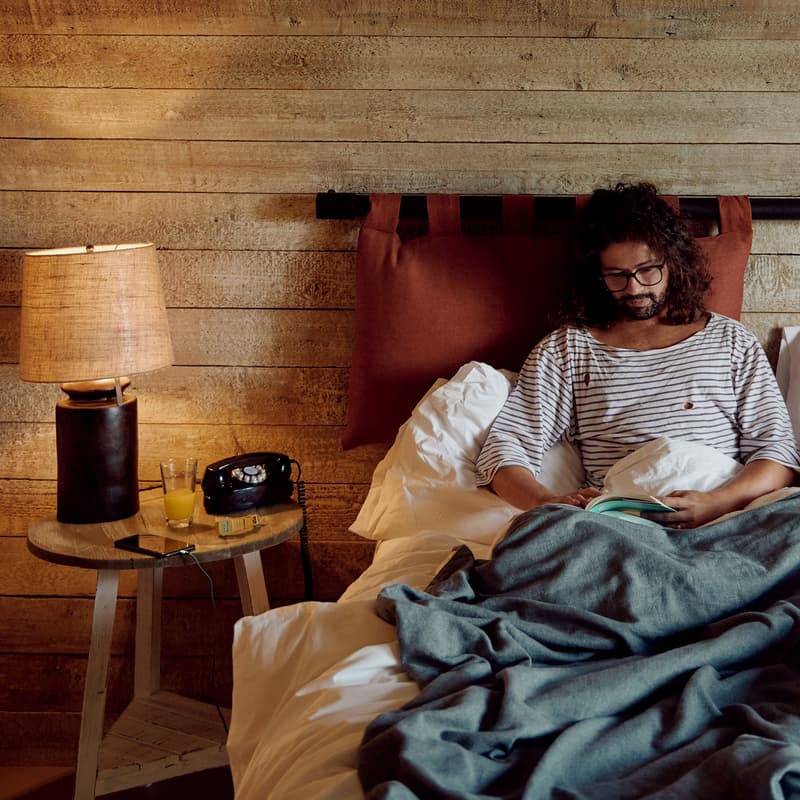 A person with curly hair and glasses reads a book in bed covered with gray and white bedding, next to a nightstand with a lamp, retro phone, drink, and smartphone. The room has rustic wooden walls.