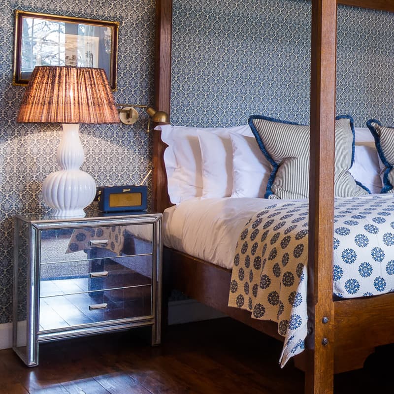 A wooden four-poster bed with blue and white bedding is set against patterned wallpaper; it is flanked by mirrored nightstands with lamps. In front, a blue velvet sofa faces a fireplace.