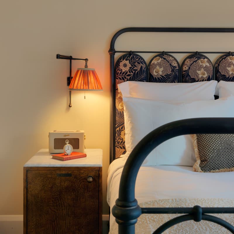 Bed with ornate headboard under a wall-mounted lamp next to a nightstand holding a retro radio, books, and an alarm clock in a cozy, softly-lit bedroom.