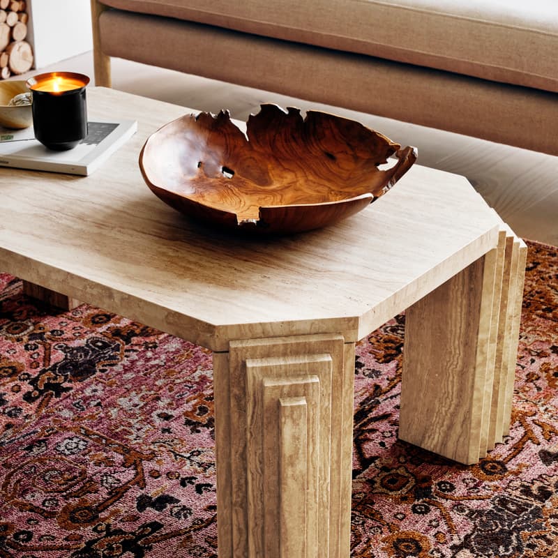 A carved wooden bowl and a black candle sit on a beige stone coffee table. Context: patterned rug, beige sofa with pillows, wood logs stacked in the background.