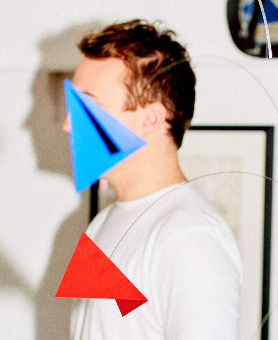 A person stands indoors, partially obscured by geometric shapes (blue and red triangles), with a background of bookshelves and framed artworks on the walls.