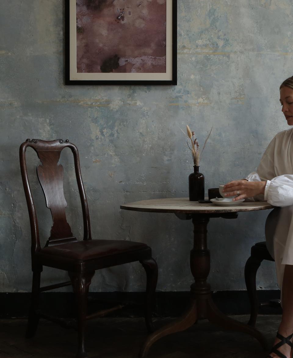 Woman wearing a white dress sits at a small round table, holding a teacup in a rustic cafe with wooden chairs, a floral picture, and a faded wall.