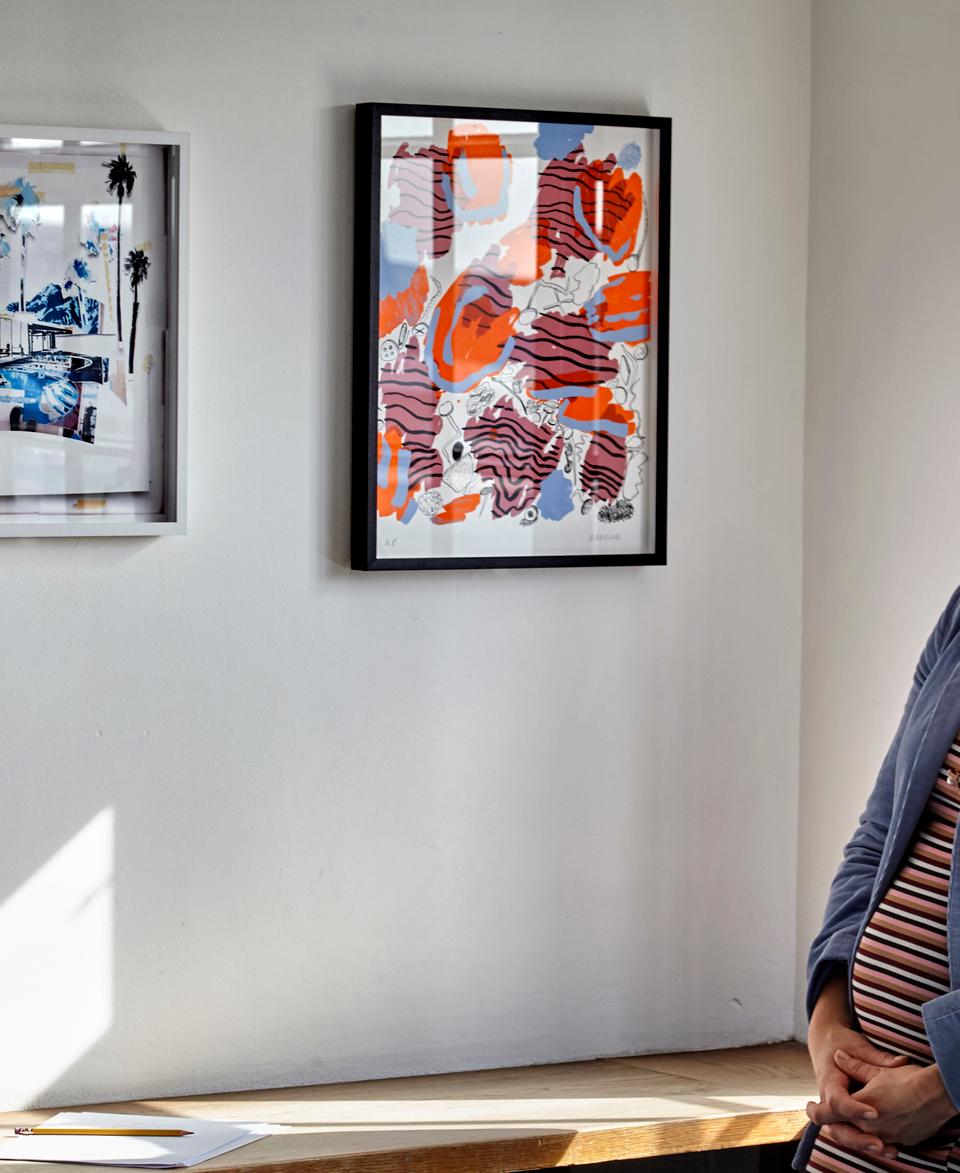 A woman with long hair, wearing a blue coat, stands gazing at two framed artworks on a white wall in a sunlit gallery space.