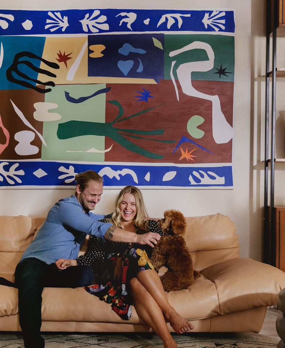 A couple on a beige sofa joyfully plays with a small brown dog in a living room adorned with a colorful abstract painting, a wooden shelf with ceramics, and candles.