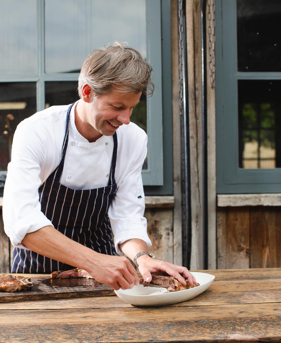 A chef is preparing food in a rustic outdoor setting, slicing meat on a wooden table in front of a wooden building with large gray-framed windows.