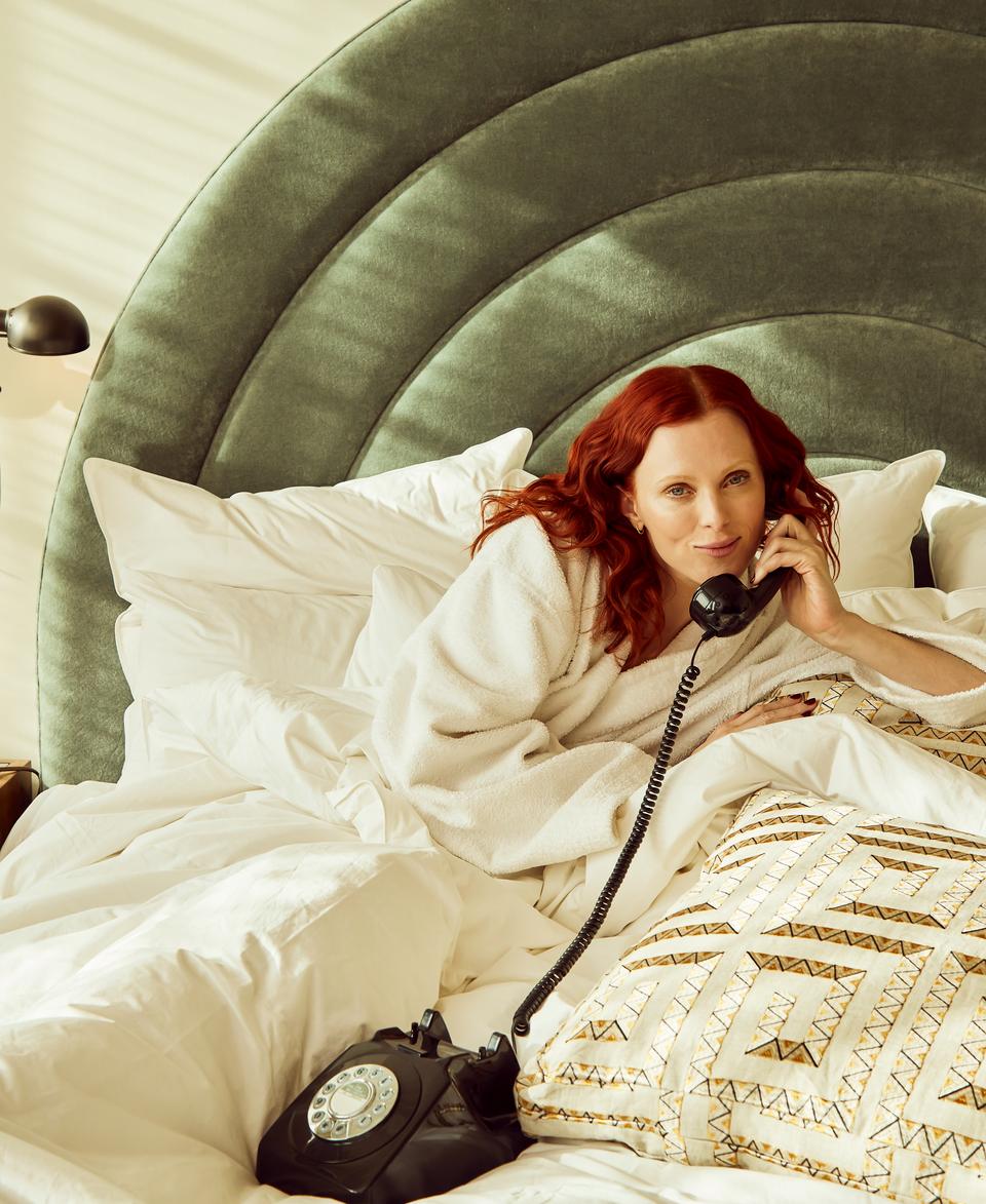 A woman, wearing a white bathrobe, lies on a bed talking on a rotary phone. The bed is adorned with white bedding and patterned pillows; a lamp sits on a wooden bedside table.
