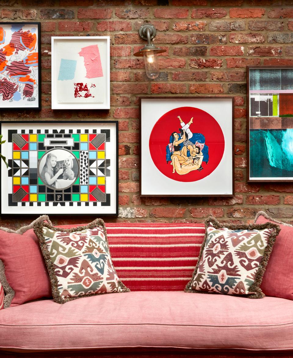 A red couch with patterned pillows sits against a brick wall displaying various framed artworks and a potted plant to the left.