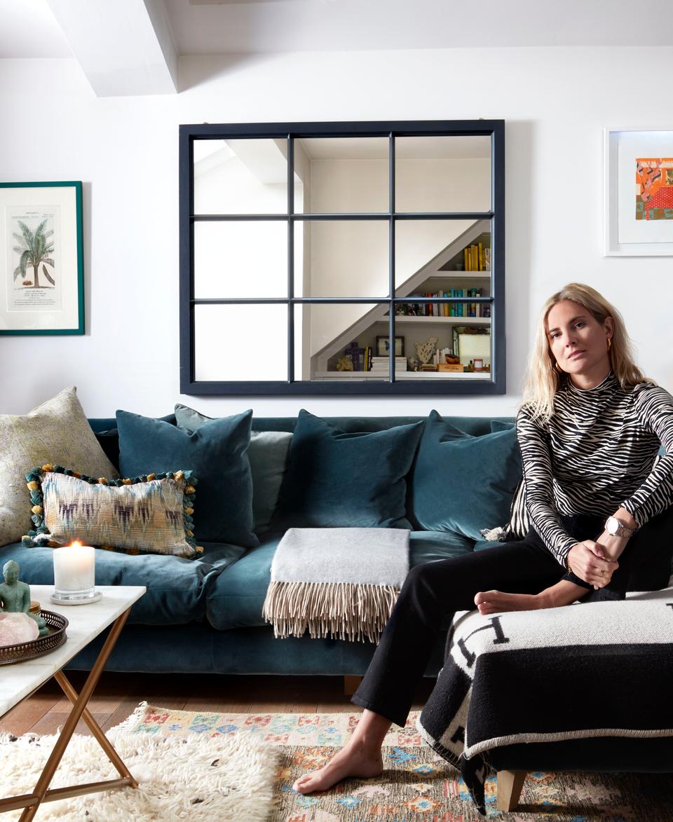 Woman sitting on a gray chaise in a stylish, modern living room with white walls, blue sofa, framed artwork, books, plants, a coffee table with books and candles, and a bar cart.