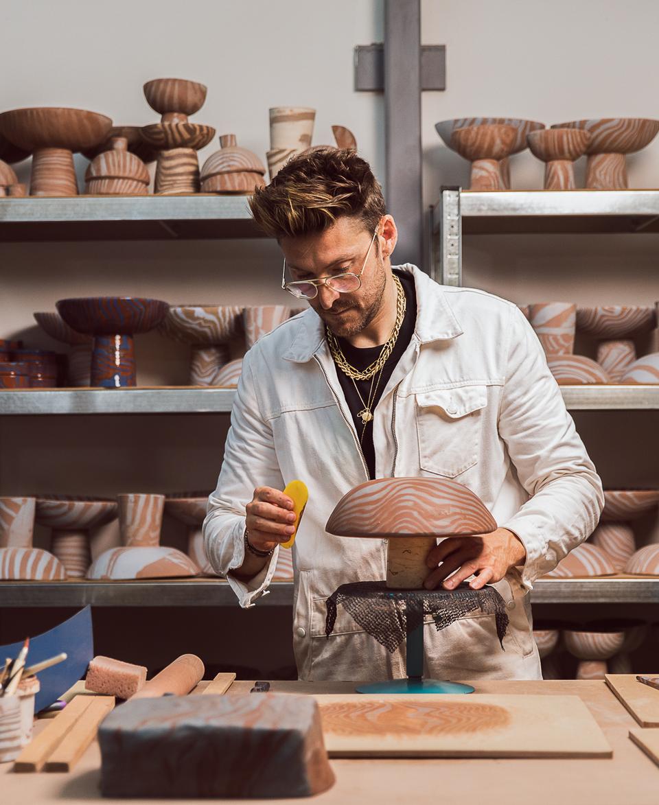 A person meticulously handcrafts a wooden bowl, using a tool for detailing, in a workshop surrounded by various shaped and patterned wooden bowls on metal shelves and a cluttered workbench.