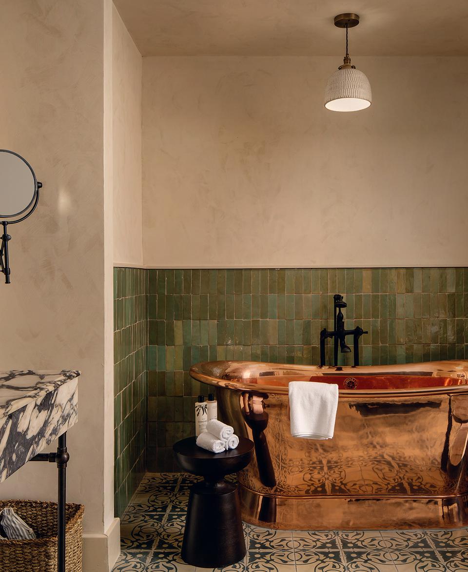 Copper bathtub with a towel hanging over it, set against green tiled walls. Marble sink with products, mirror, and magnifying mirror. Black stool with rolled towels and a plant.