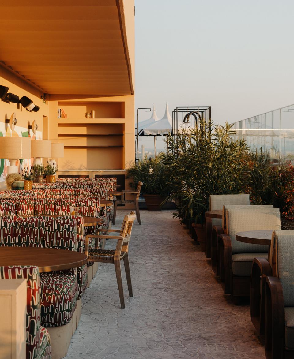 Rooftop restaurant features patterned chairs and round wooden tables set along a decorative wall with lamps and plants, offering an airy and lush ambiance with city views.