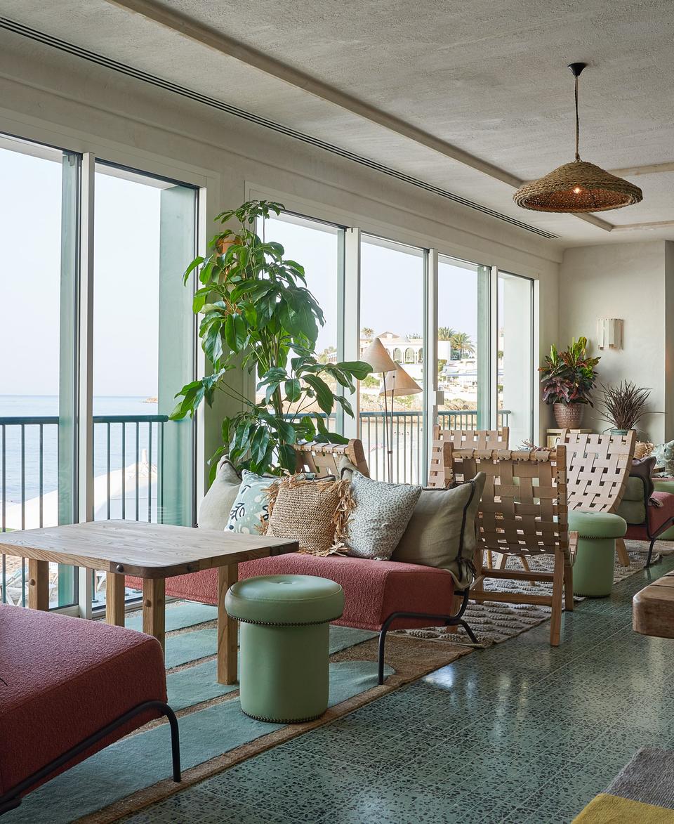 Seating area with cushions and small tables, overlooking the ocean through large windows. Indoor plants are scattered around, and pendant lights hang from the ceiling in a cozy lounge setting.