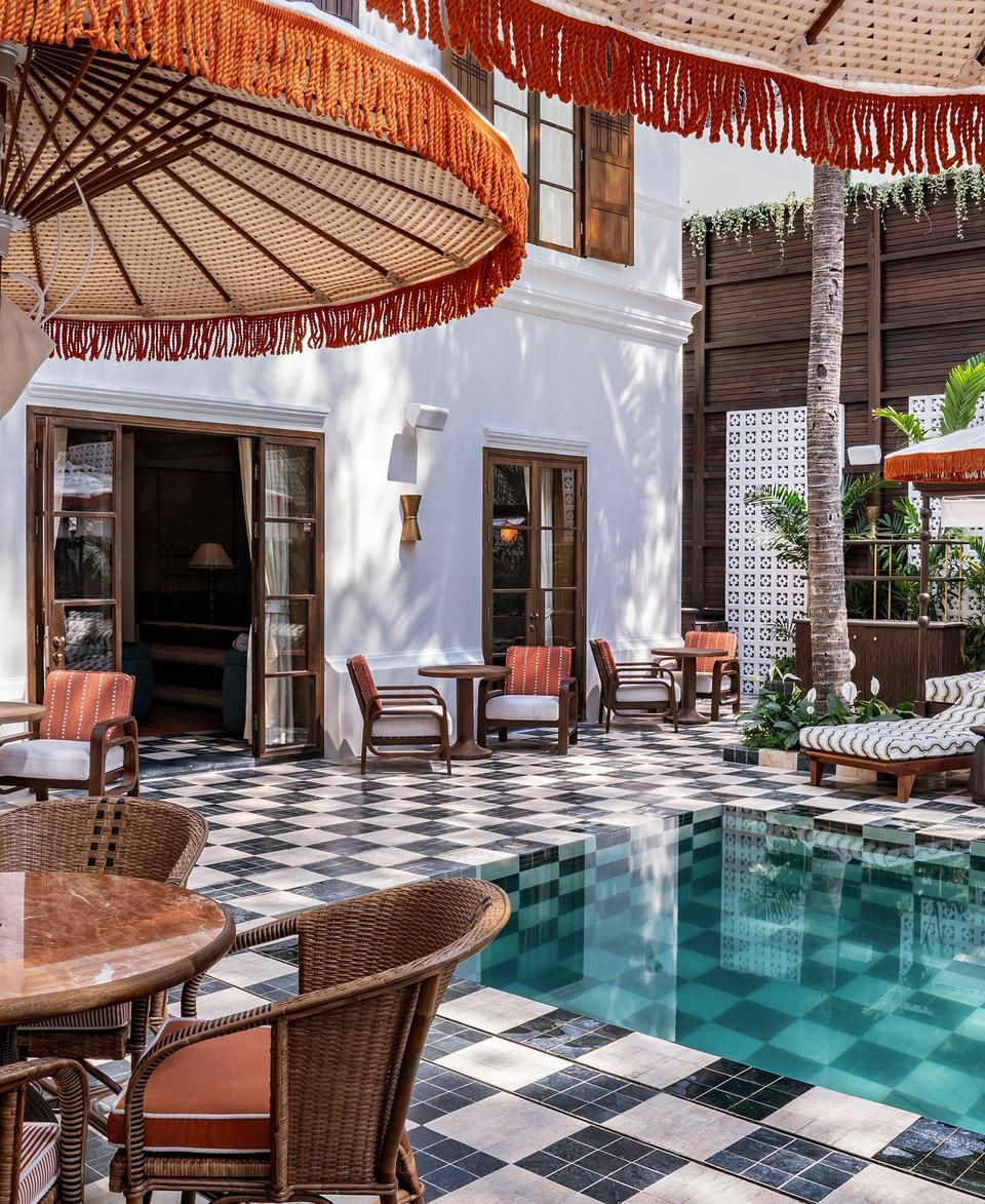 Poolside lounge chairs with umbrellas surround an empty, turquoise-tiled swimming pool. The area, bordered by tropical plants and a white building with wooden doors and windows, features wicker furniture and checkered flooring.