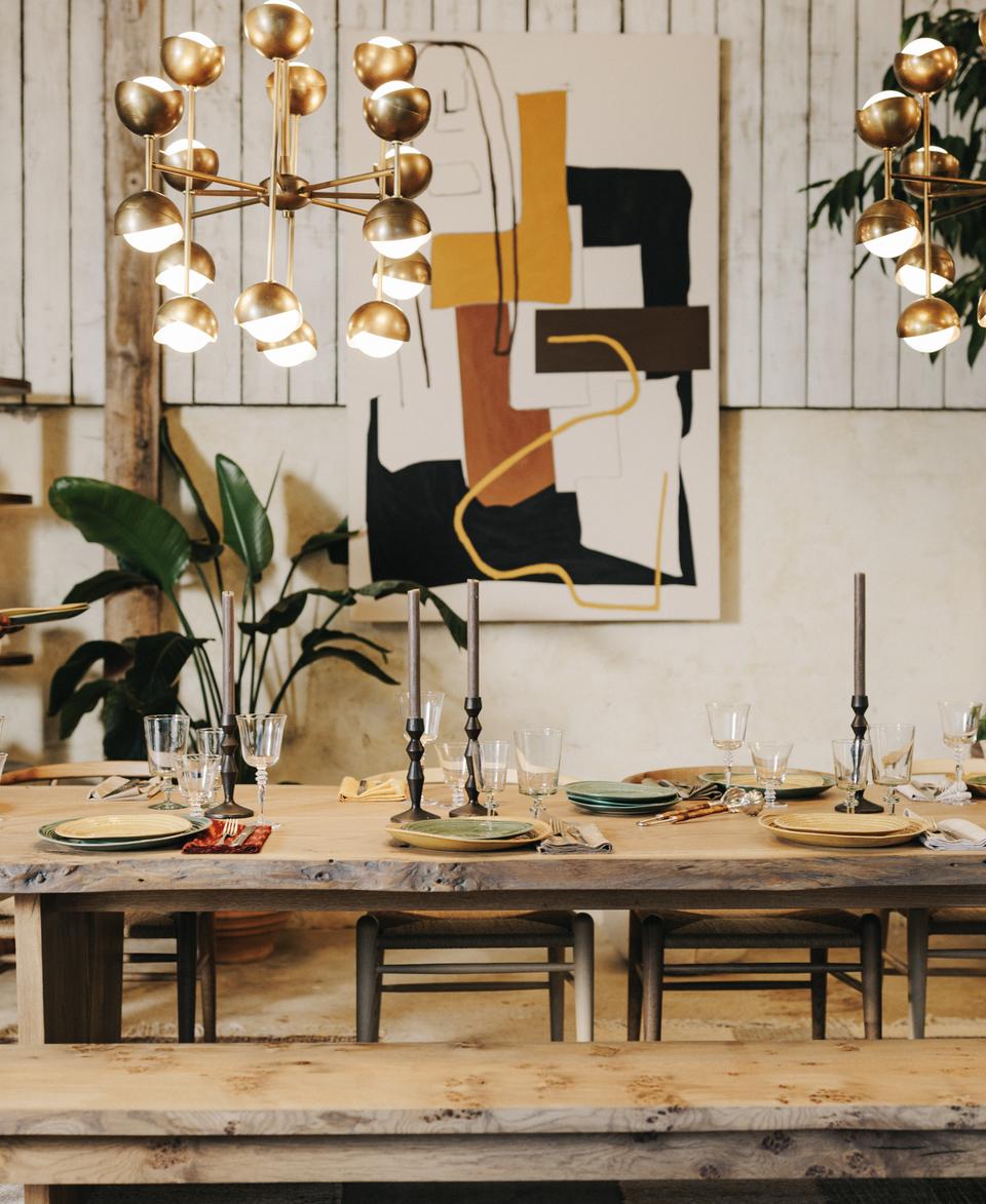 A woman in a light blue sweater is setting a dining table with plates and glassware in a rustic, well-decorated room with plants, shelves, abstract art, and gold chandeliers.