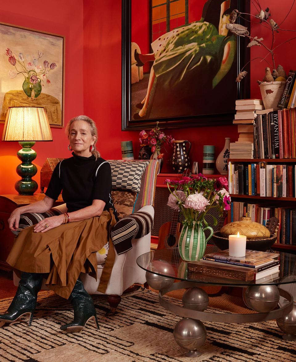A woman sits on a patterned sofa in a vibrant red-walled living room surrounded by bookshelves, art, flowers, a green lamp, and decorative objects. A striped rug covers the floor.