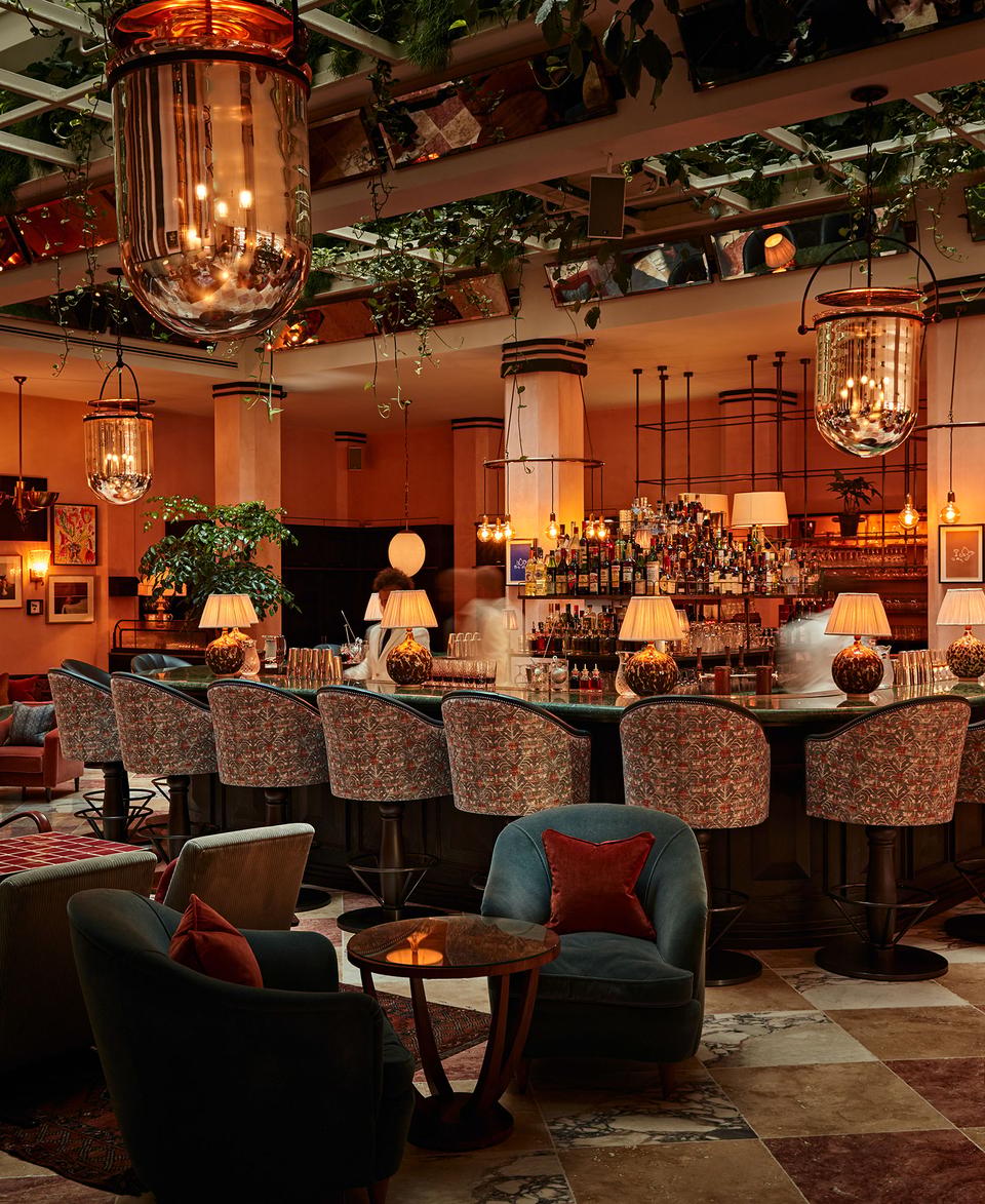 A large, elegant bar with cushioned, patterned stools, illuminated by hanging vintage lamps, surrounded by lush indoor plants, cozy armchairs, and small round tables in a warm, sophisticated setting.