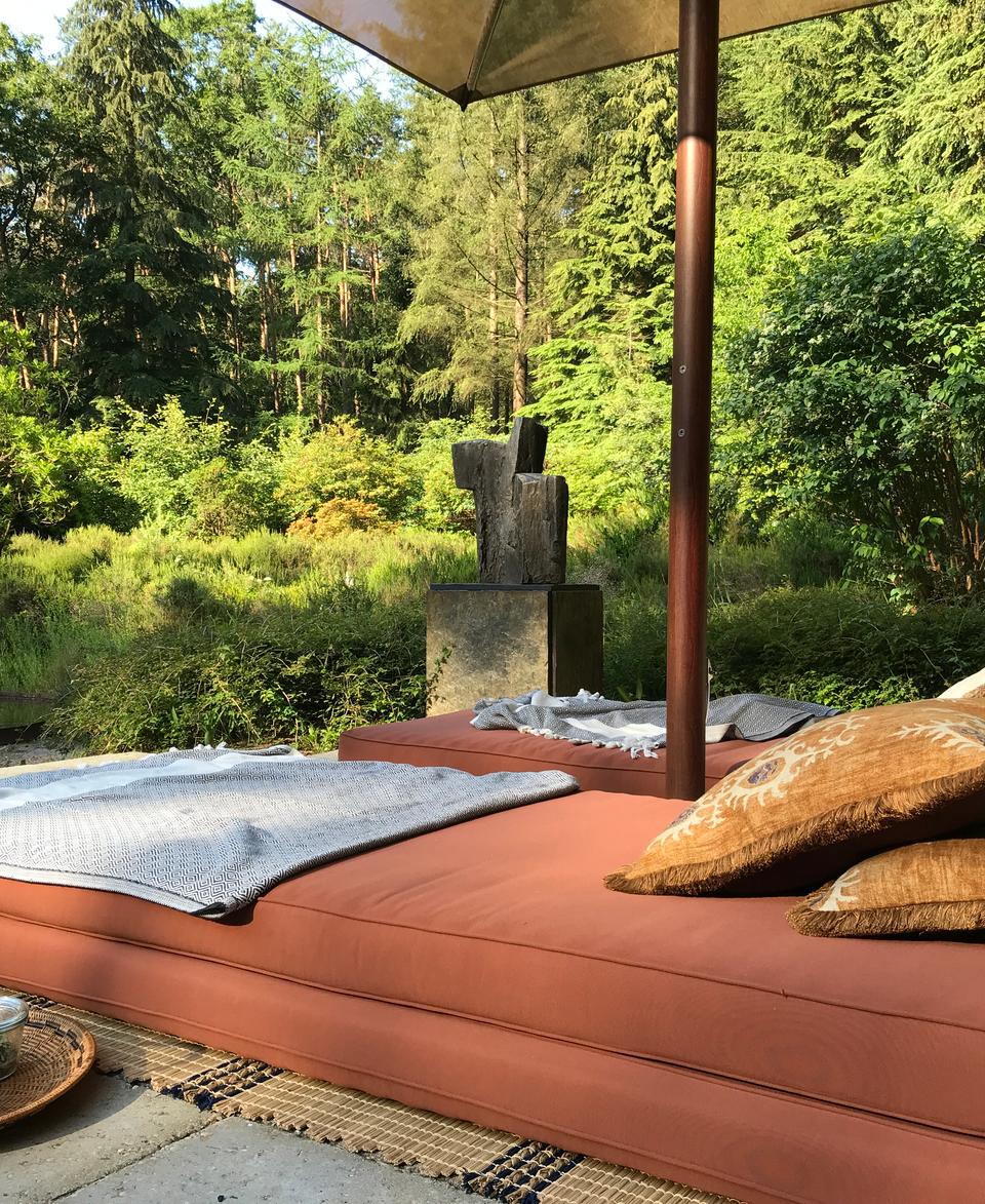 A shaded outdoor seating area features an orange cushioned daybed with pillows, a blanket, and a tea set, surrounded by lush, green vegetation and trees.
