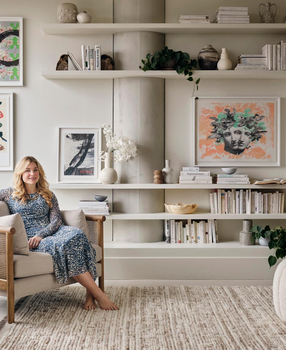 A woman in a patterned dress sits barefoot in a beige armchair in a stylish living room with art-adorned walls, bookshelves, indoor plants, and a textured rug.