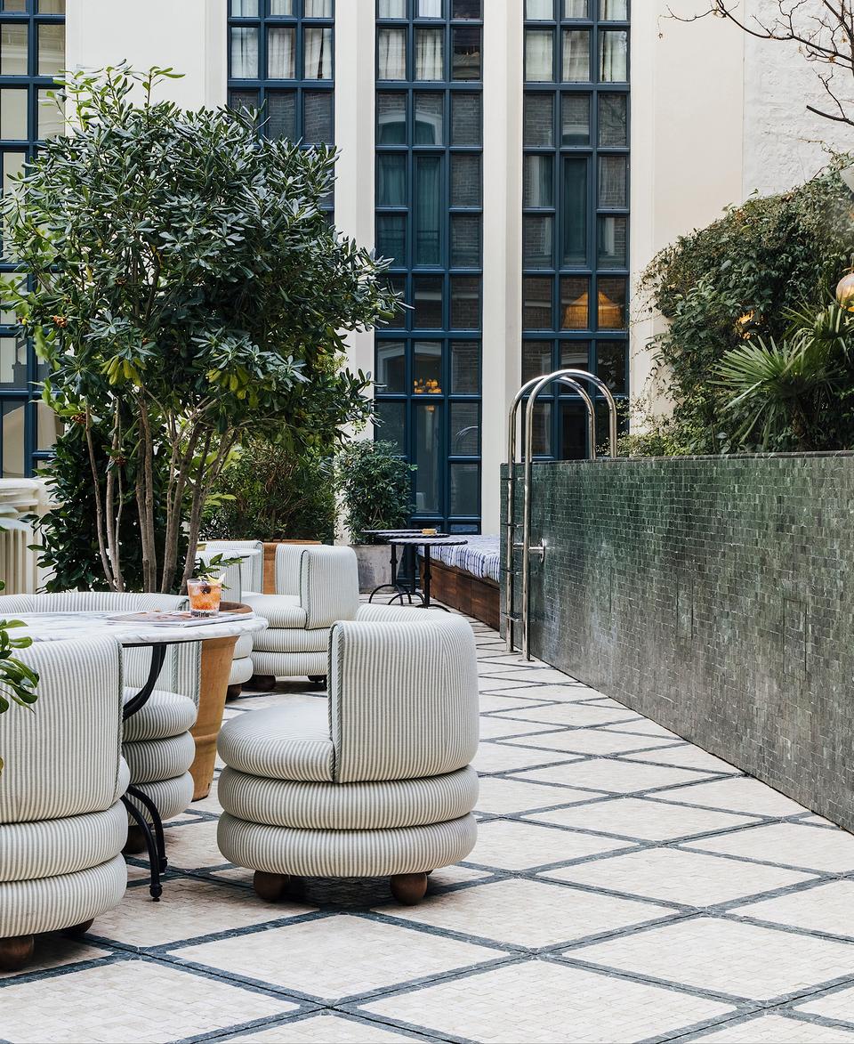 Plush seats surround a table with drinks in a stylish outdoor patio adorned with potted plants and framed by tall windows and a green-tiled wall.