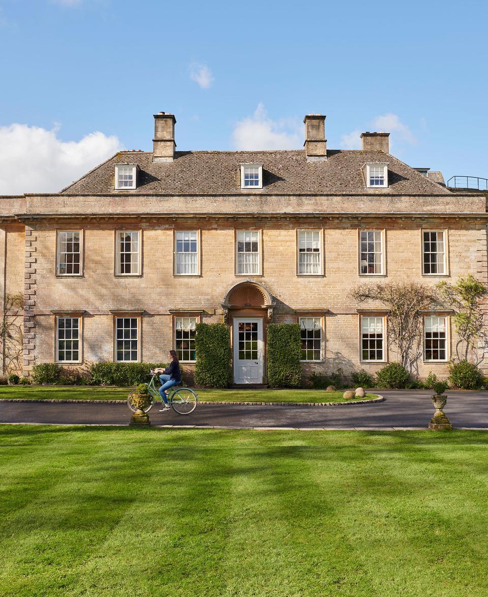 A person rides a bicycle past a large, symmetrical, three-story stone mansion with multiple windows in a landscaped garden with neatly trimmed grass and hedges under a bright blue sky.