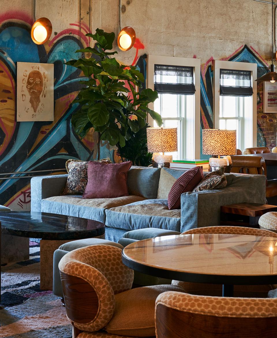 A cozy seating area with a gray sofa, patterned chairs, and round tables is in a room featuring large windows, colorful graffiti walls, hanging lights, and indoor plants.
