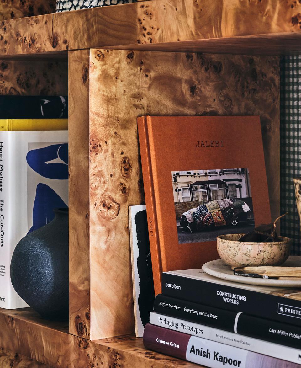 Books and decorative items sit on a wooden shelf. Books include "Henri Matisse The Cut-Outs," "Jalebi," and "Anish Kapoor." A vase, bowl, and candle form the display. The shelf is against a checkered fabric background.