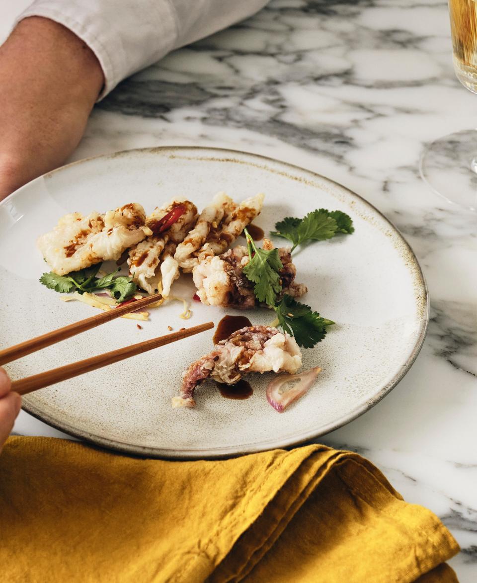 A person uses chopsticks to pick up a piece of fried food on a white plate garnished with greens, set on a marble table with a yellow napkin and a glass of white wine.