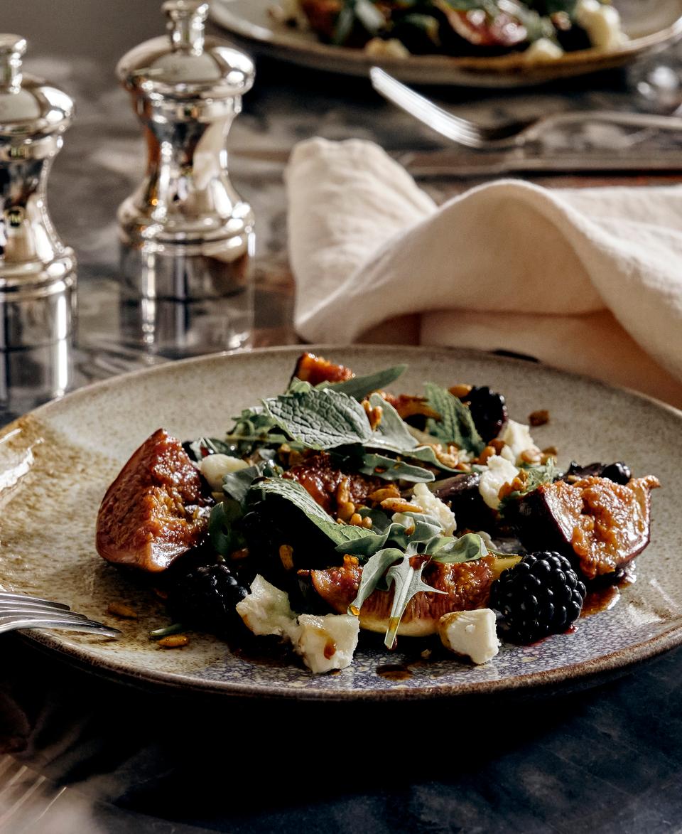 A plated salad with figs, blackberries, leafy greens, goat cheese, and nuts rests on a marble table, accompanied by a glass of red wine, cutlery, two metal shakers, and a cloth napkin.