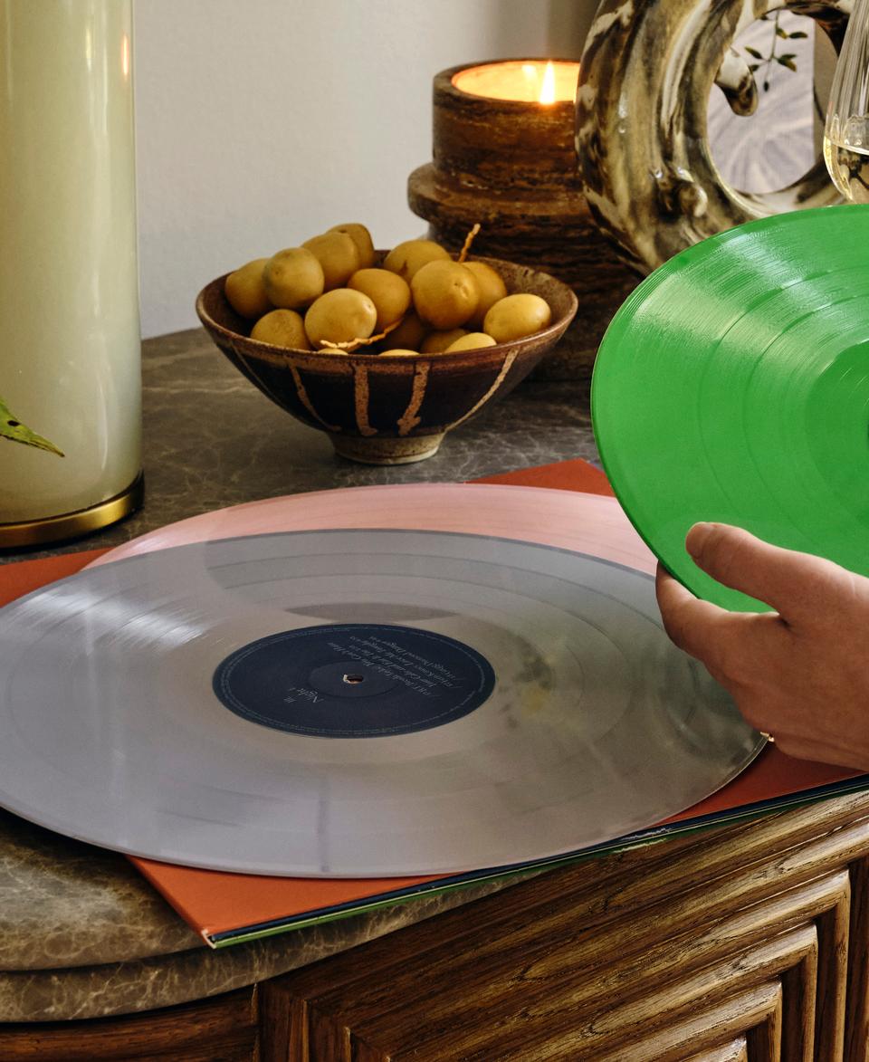 A hand holds a green vinyl record above a table. Another vinyl record lies on the table. Nearby are a candle, a fruit bowl, a plant, and a glass of white wine.